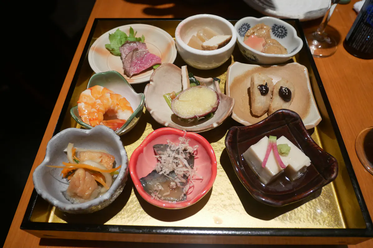 Japanese Cuisine in Kyoto A wooden tray holding an assortment of small Japanese dishes, including tofu, fish, shrimp, pickled vegetables, and garnishes, arranged in various bowls and plates. Each dish is artfully presented, showcasing a variety of colors and textures.
