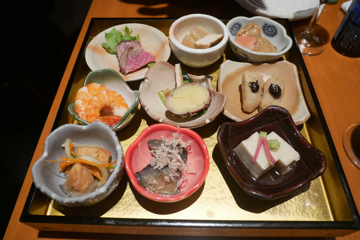 A wooden tray holding an assortment of small Japanese dishes, including tofu, fish, shrimp, pickled vegetables, and garnishes, arranged in various bowls and plates. Each dish is artfully presented, showcasing a variety of colors and textures.