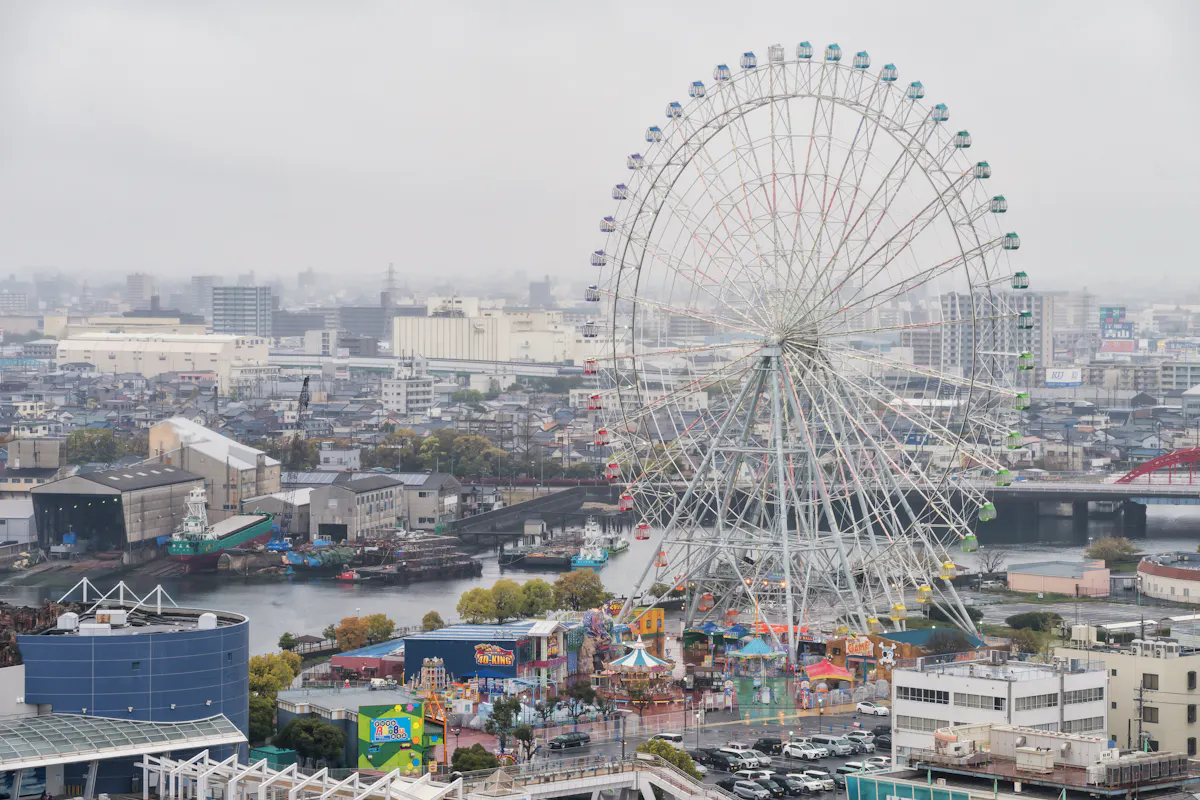 Port of Nagoya An aerial view of a cityscape featuring a large Ferris wheel with multicolored cabins. The surrounding area includes various buildings, a river with boats, and a colorful amusement park with several attractions. The sky is overcast, creating a misty atmosphere.