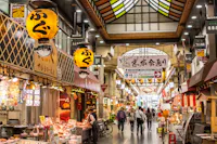 Indoor Japanese market with hanging lanterns, various food stalls, and people walking down the aisle. Colorful signs and banners hang from the ceiling, creating a lively, bustling atmosphere.