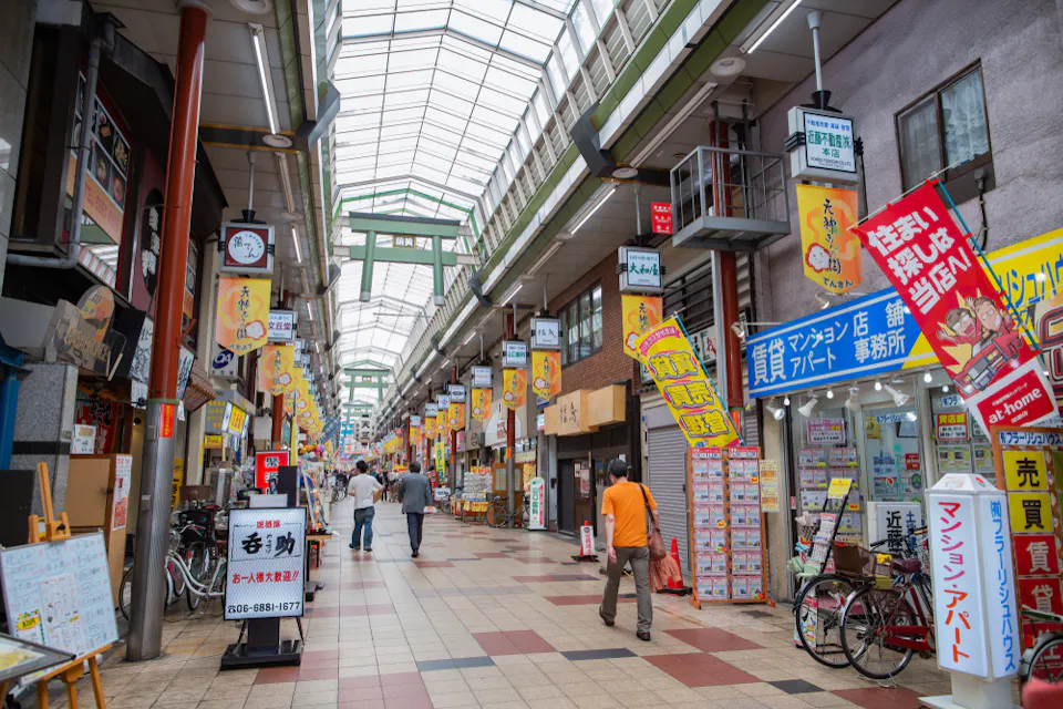 A covered shopping arcade in Japan with various colorful signs and advertisements in Japanese. Pedestrians walk through, while bicycles are parked along the sides. The arcade has a glass ceiling and a tiled floor.