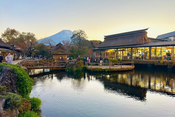 A scenic view of traditional Japanese buildings by a pond at sunset, with people walking on wooden bridges and Mount Fuji visible in the background.