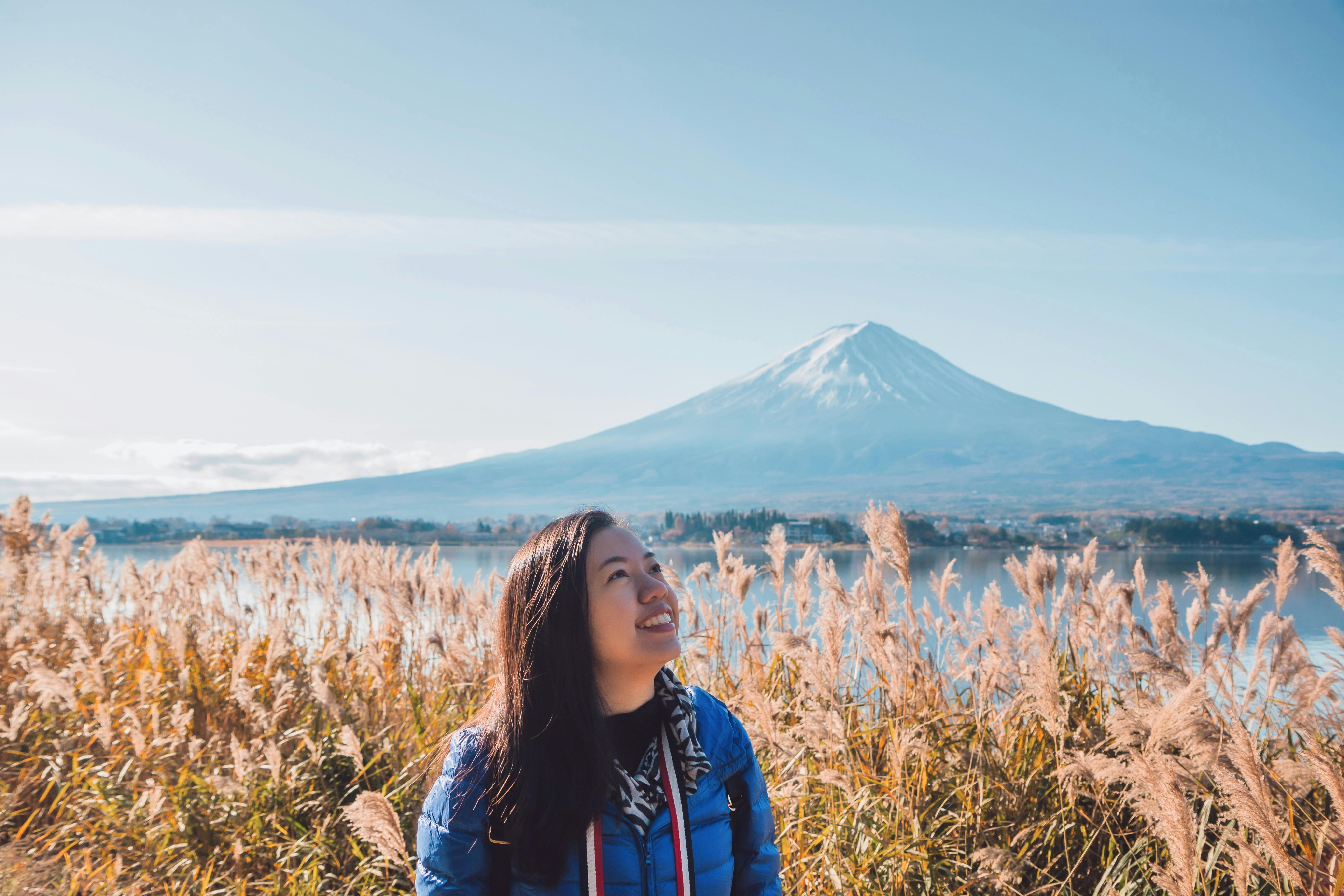 A woman in a blue jacket smiles while standing in tall grass, with Mount Fuji and a clear blue sky in the background.