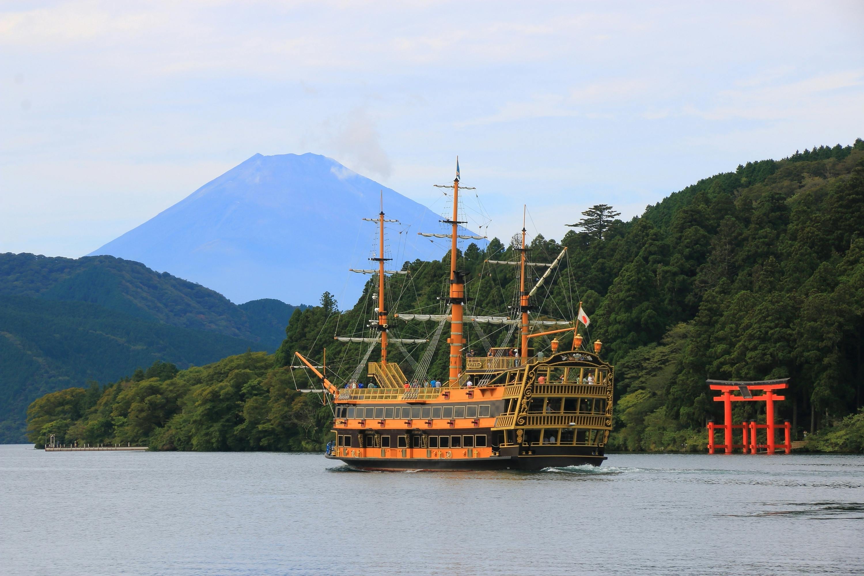 A large, yellow and black pirate-style ship sails on a lake surrounded by green trees, with Mount Fuji in the background and a red torii gate near the shoreline.