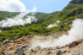 Steam rises from rocky ground in a lush, green mountainous area under a bright blue sky, suggesting geothermal activity in a volcanic landscape.
