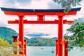 A bright red torii gate stands in the water near a lake, surrounded by green trees and hills under a partly cloudy sky. A small boat floats on the lake in the background.