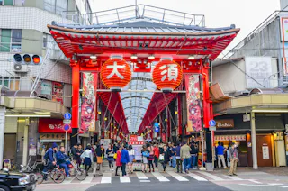 A bustling marketplace entrance adorned with large red lanterns and traditional signage, with people walking and shopping. The covered walkway is surrounded by shops and buildings on either side under a red and white roof structure.