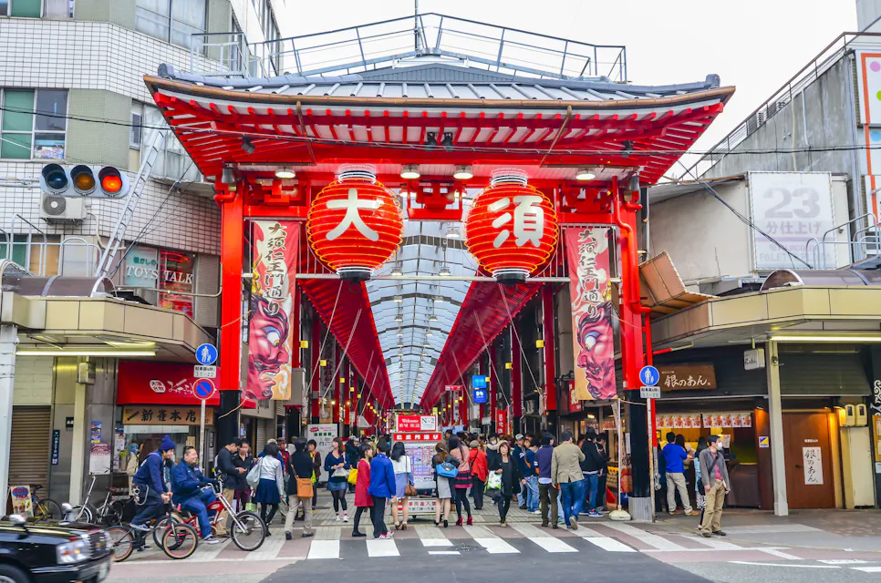 A bustling marketplace entrance adorned with large red lanterns and traditional signage, with people walking and shopping. The covered walkway is surrounded by shops and buildings on either side under a red and white roof structure.
