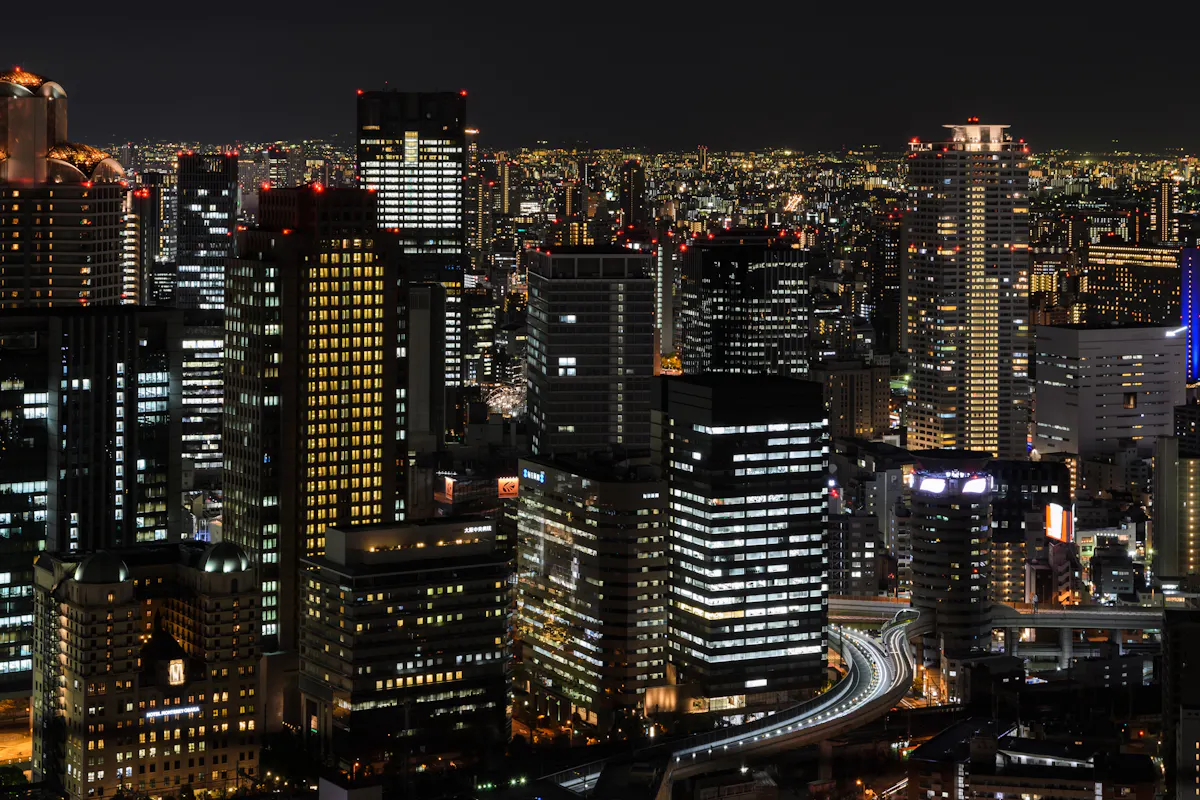 Umeda Sky Building in Osaka A nighttime cityscape featuring numerous illuminated high-rise buildings, creating a stunning array of lights. Roads and highways weave through the buildings, and the skyline is bathed in the soft glow of city lights under a dark sky.