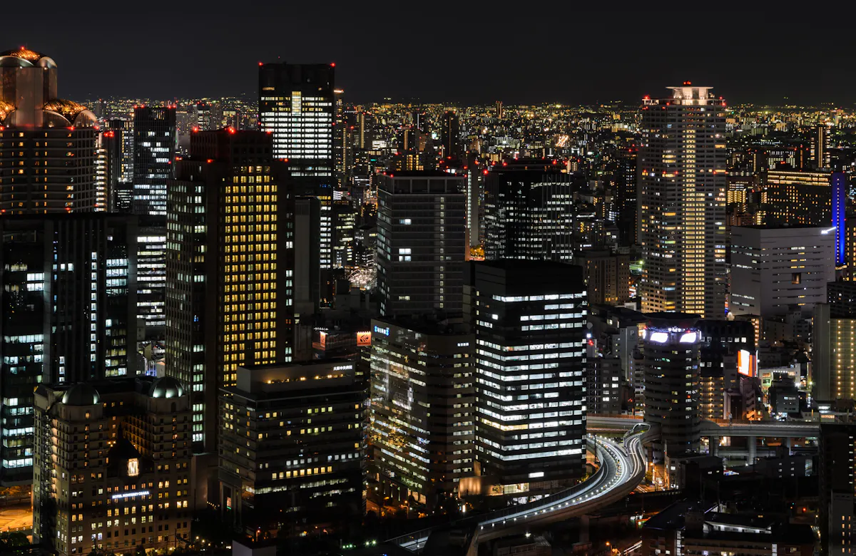 A nighttime cityscape featuring numerous illuminated high-rise buildings, creating a stunning array of lights. Roads and highways weave through the buildings, and the skyline is bathed in the soft glow of city lights under a dark sky.