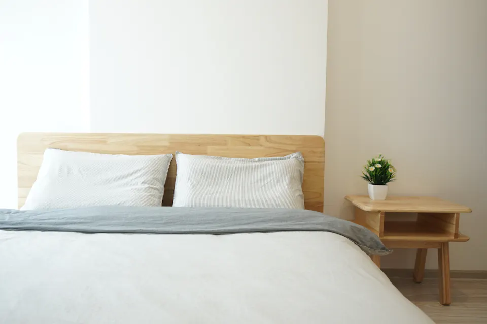 一个极简主义的卧室，配有木制床架和床头板。床上铺着白色床单和两个枕头，上面覆盖着一条浅灰色的毯子。床旁是一个木制床头柜，放着一盆小植物。墙壁刷成白色。