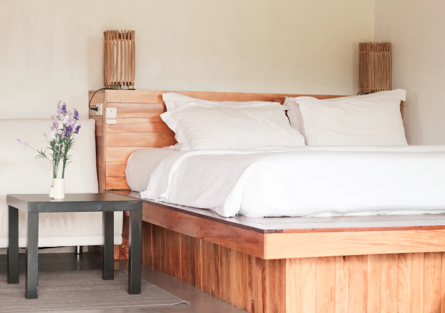 一个舒适的卧室，配有木制床架，白色床品和枕头。床旁是一个小黑色咖啡桌，上面摆放着一瓶紫色花朵。房间设计简约，柔和的光线和米色墙壁相得益彰。