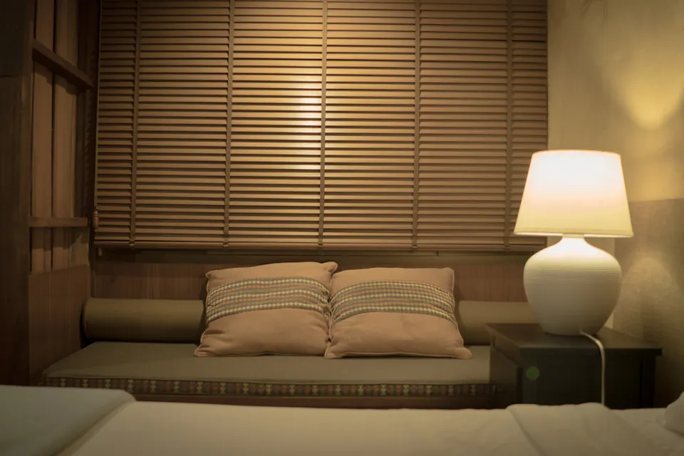 一个舒适的角落，配有一个软垫长椅和两个棕色靠垫，背靠着木百叶窗。一盏温暖的白色台灯放在深色边桌上，柔和地照亮空间，营造出宁静宜人的气氛。在前景中可以看到一张铺着白色床上用品的床边缘。