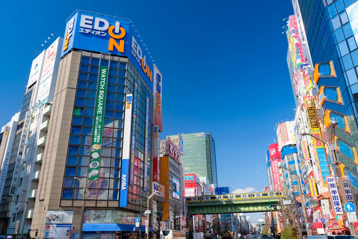Colorful city street in Japan with tall buildings covered in bright signs and advertisements, a train passing on an elevated track, and a clear blue sky above.