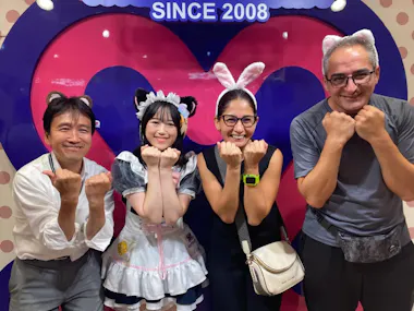 Four people pose in front of a heart-shaped sign, wearing animal ear headbands and making playful paw gestures. One person is dressed in a maid costume, the others are casually dressed and smiling.