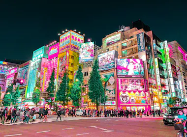 A bustling city street at night in Tokyo, Japan, with crowds crossing the road and colorful neon signs and billboards illuminating tall buildings lining the intersection.