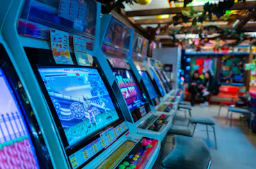 A row of colorful arcade game machines with screens and controls, standing side by side in a retro gaming arcade. A few people are seated in the background, slightly out of focus.