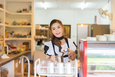 A smiling barista in a black and white uniform stands behind a counter in a coffee shop, holding a metal pitcher and preparing a drink. Cups and coffee equipment are visible around her.