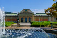 A historic building with a dome roof and ornate facade is surrounded by lush greenery under a clear blue sky. In the foreground, a fountain with arched water jets adds a dynamic element to the serene landscape.