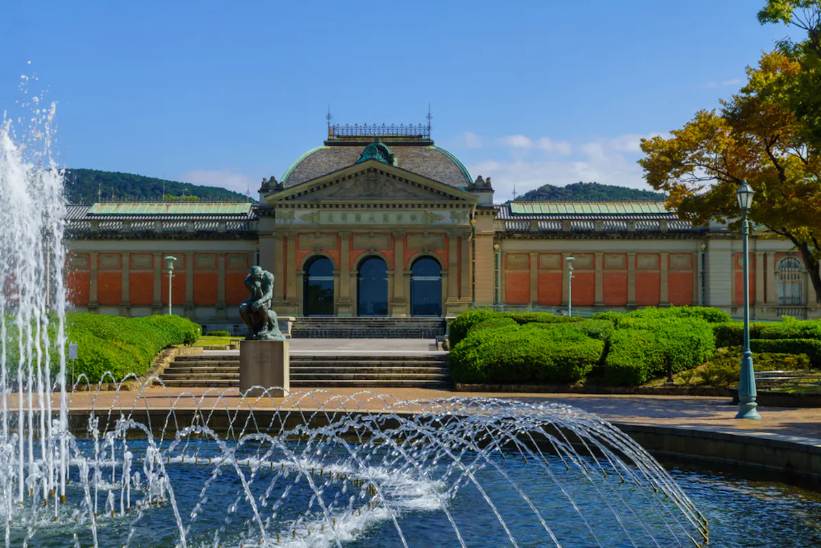 A historic building with a dome roof and ornate facade is surrounded by lush greenery under a clear blue sky. In the foreground, a fountain with arched water jets adds a dynamic element to the serene landscape.