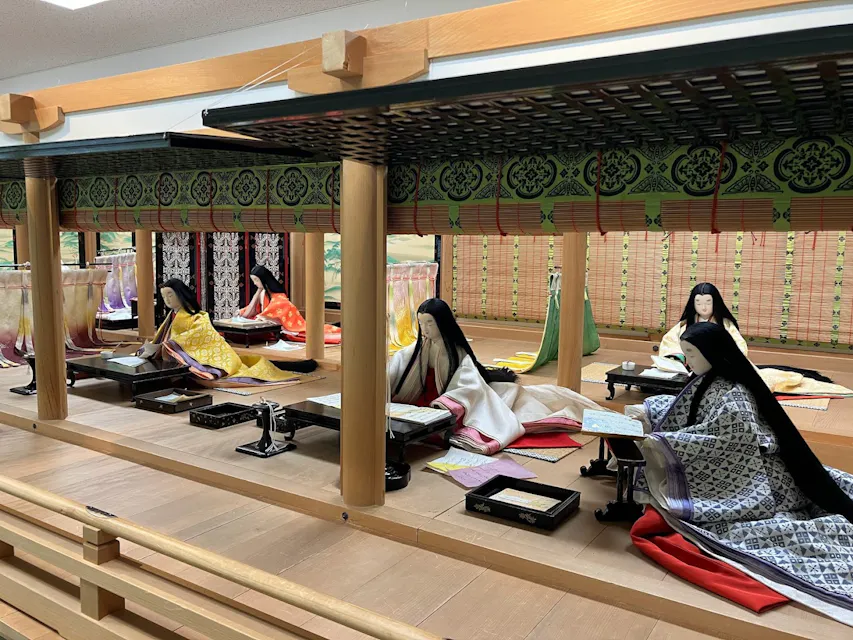 伝統的な平安時代の衣装を身にまとった日本の宮廷人形の詳細な展示で、低いテーブルの周りに様々なミニチュアアイテムが置かれています。シーンは飾られた緑の天蓋と背景に模様のあるカーテンがある木造の構造物の中に設定されています。