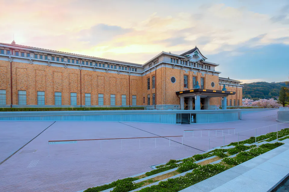 夕暮れ時に映し出された、淡いオレンジ色のレンガ外壁と華麗な日本の建築要素を持つ伝統的な建物。入り口には金のアクセントが施された黒いオーバーハングがあり、前景には舗装されたエリアと手入れの行き届いた緑が広がっている。