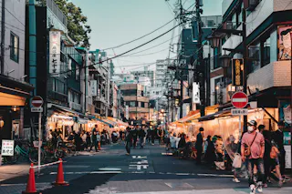 Busy street market in Japan with people walking and shopping. Outdoor food stalls line both sides, signs with Japanese characters hang above, and overhead power lines crisscross the sky. Some people wear face masks.