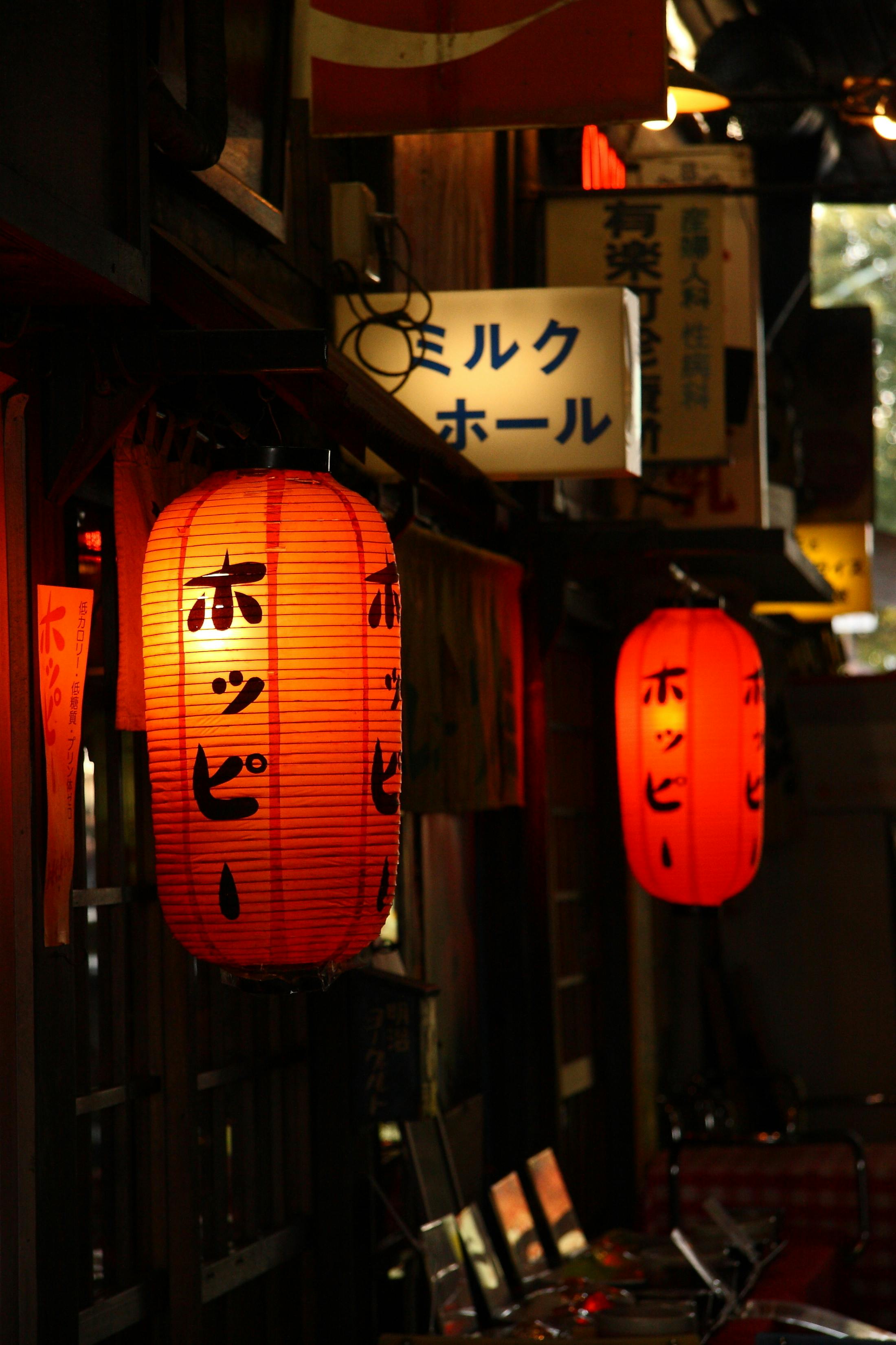 A dimly lit street scene with glowing red Japanese lanterns hanging outside a building, featuring black vertical Japanese writing. Signs with Japanese text are visible in the background above the entrance.