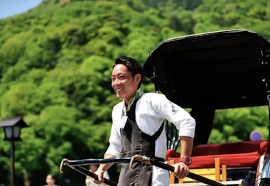 A smiling man stands outdoors next to a traditional rickshaw, holding its handles. He is wearing a white shirt and a black apron, with lush green trees and a hill in the background.
