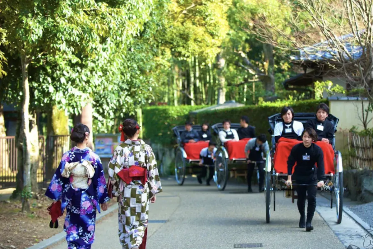 Rickshaw Ride People stroll along a tree-lined path, with some riding in rickshaws. Two women in kimonos walk side by side, while others enjoy the scenic surroundings. The setting appears tranquil and lush with greenery.