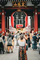 A crowd of people gathers in front of the large red lantern at the entrance of Senso-ji Temple in Tokyo, Japan, with traditional Japanese architecture and signs visible above.
