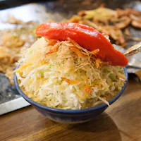 Monjayaki A bowl of fresh coleslaw topped with crispy, thinly sliced onions and a halved red bell pepper, served with a silver spoon. The background shows a flat griddle with scattered cooked vegetables.