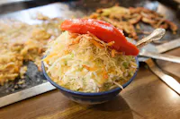 A bowl of fresh coleslaw topped with crispy, thinly sliced onions and a halved red bell pepper, served with a silver spoon. The background shows a flat griddle with scattered cooked vegetables.