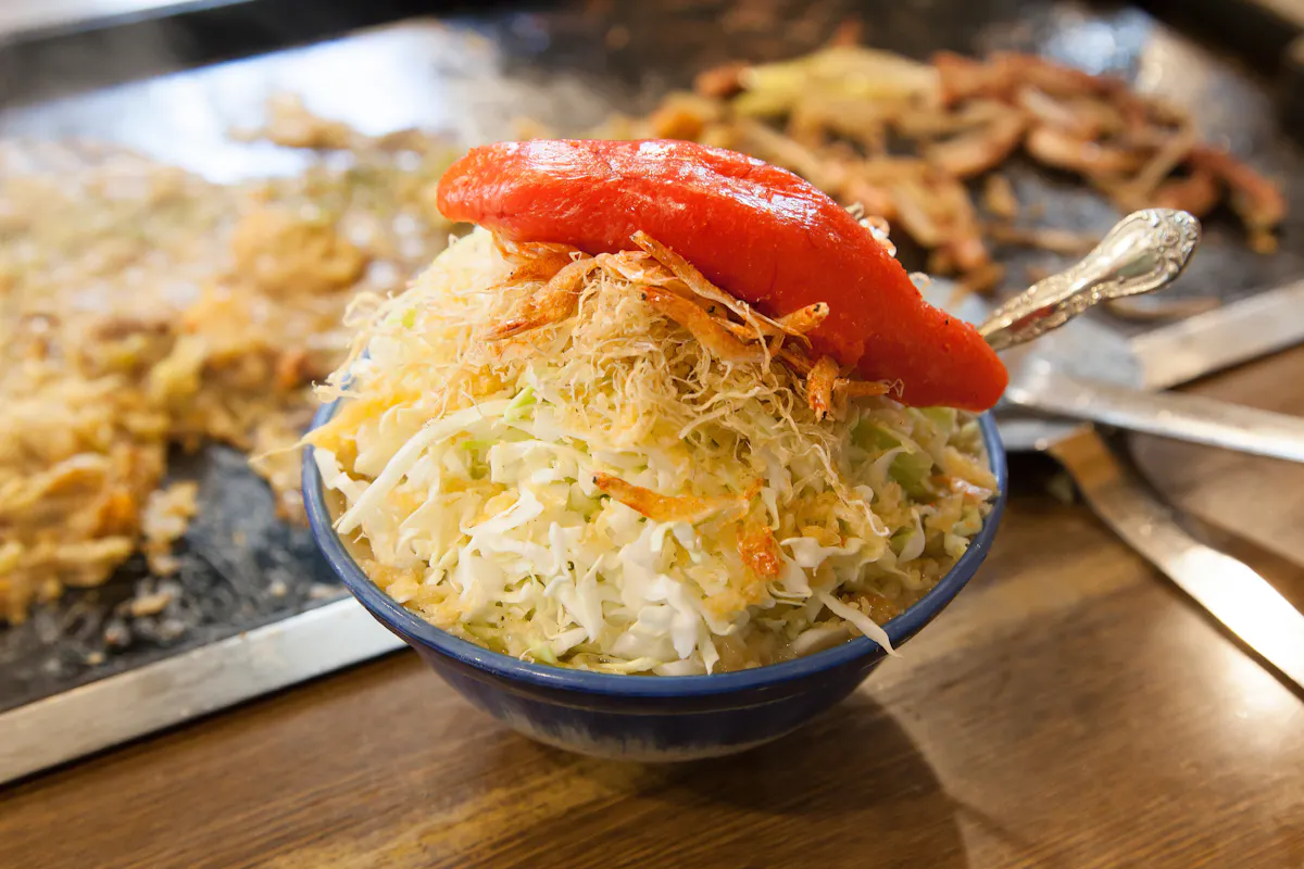A bowl of fresh coleslaw topped with crispy, thinly sliced onions and a halved red bell pepper, served with a silver spoon. The background shows a flat griddle with scattered cooked vegetables.