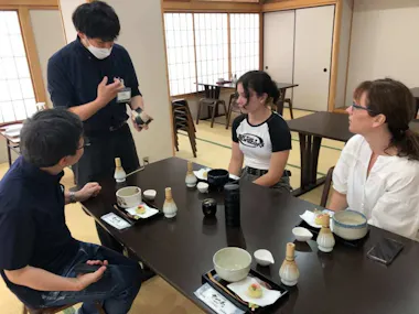 Three people sit at a table in a traditional Japanese room, listening to a man in a mask explain something. Dishes and drinks are on the table, and the setting appears calm and instructional.