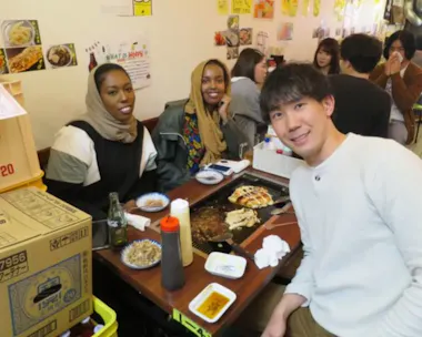 Three people sit at a restaurant table with food, drinks, and condiments. Two women wearing hijabs smile at the camera, and a man sits beside them. Other diners fill tables in the busy restaurant.