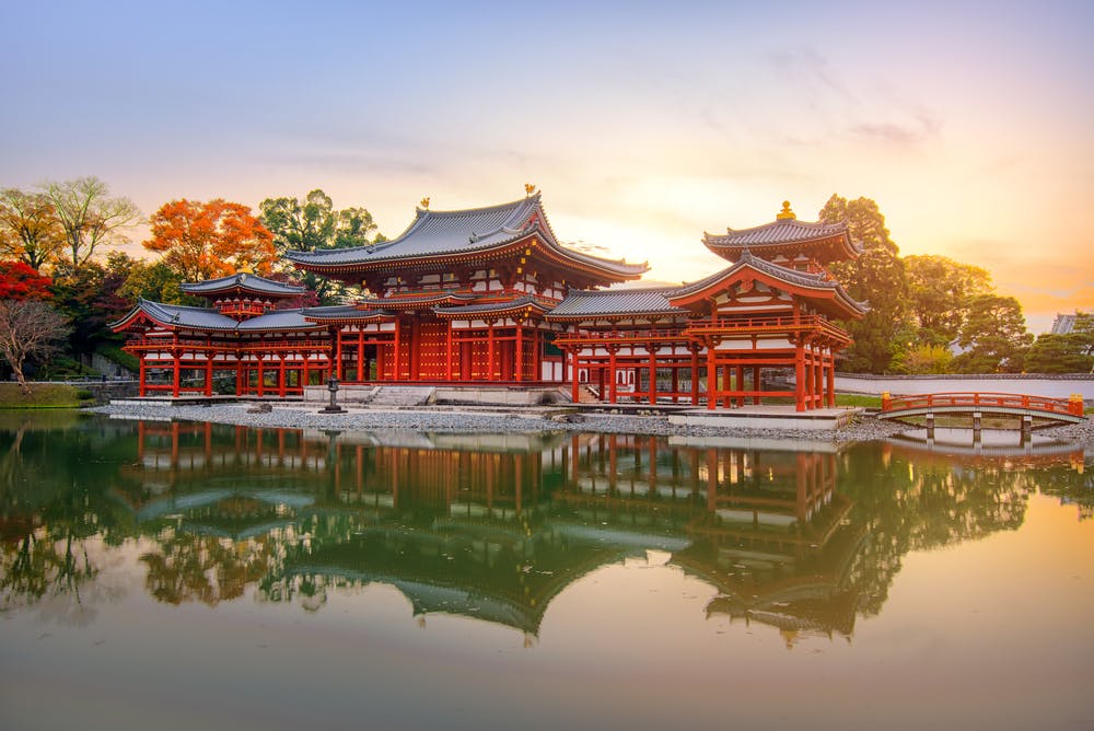 A traditional Japanese temple with ornate red and white architecture is reflected in a calm pond at sunset, surrounded by vibrant autumn trees and a small arched bridge.