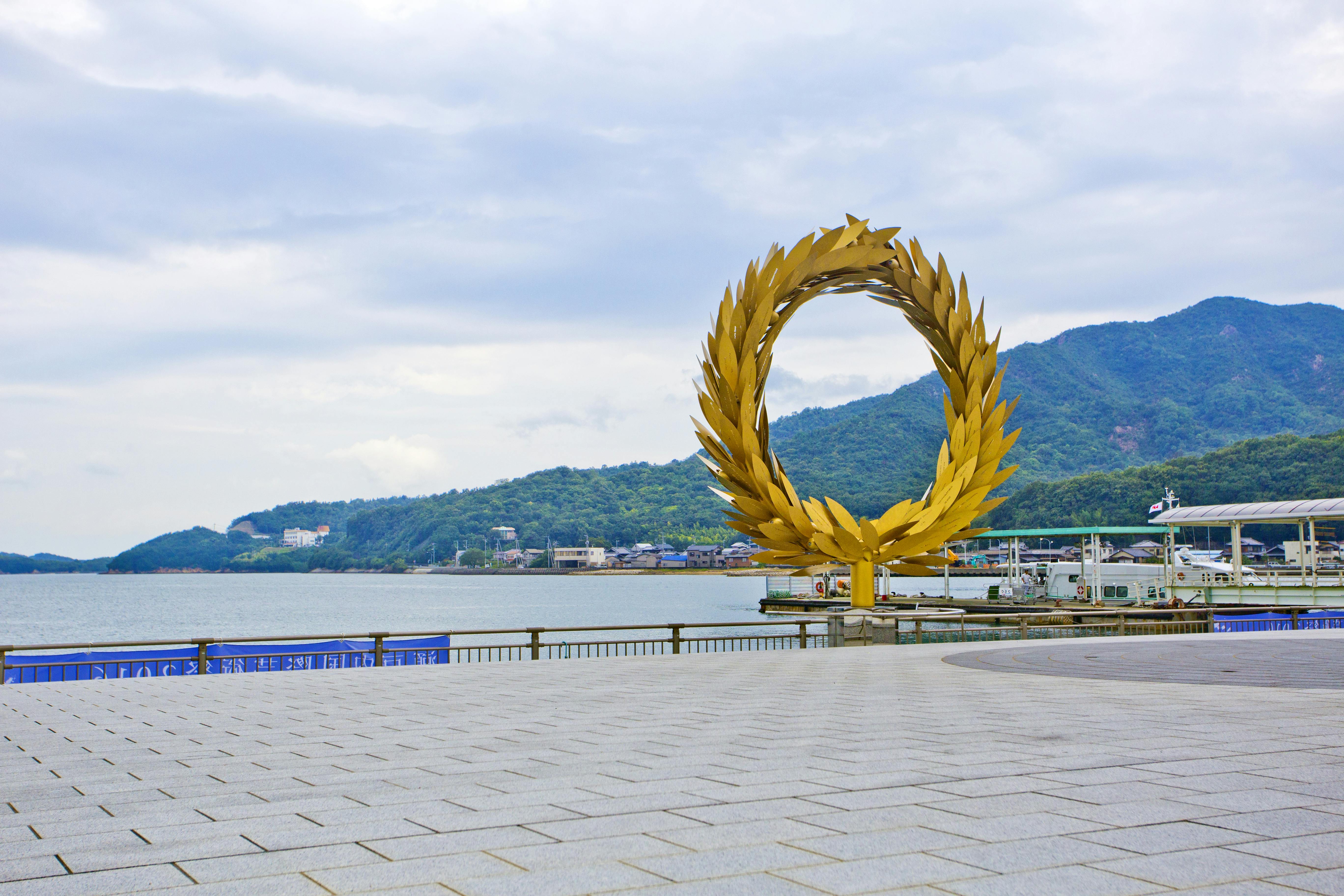 A large golden wreath sculpture stands near a waterfront promenade with mountains and boats in the background under a cloudy sky.
