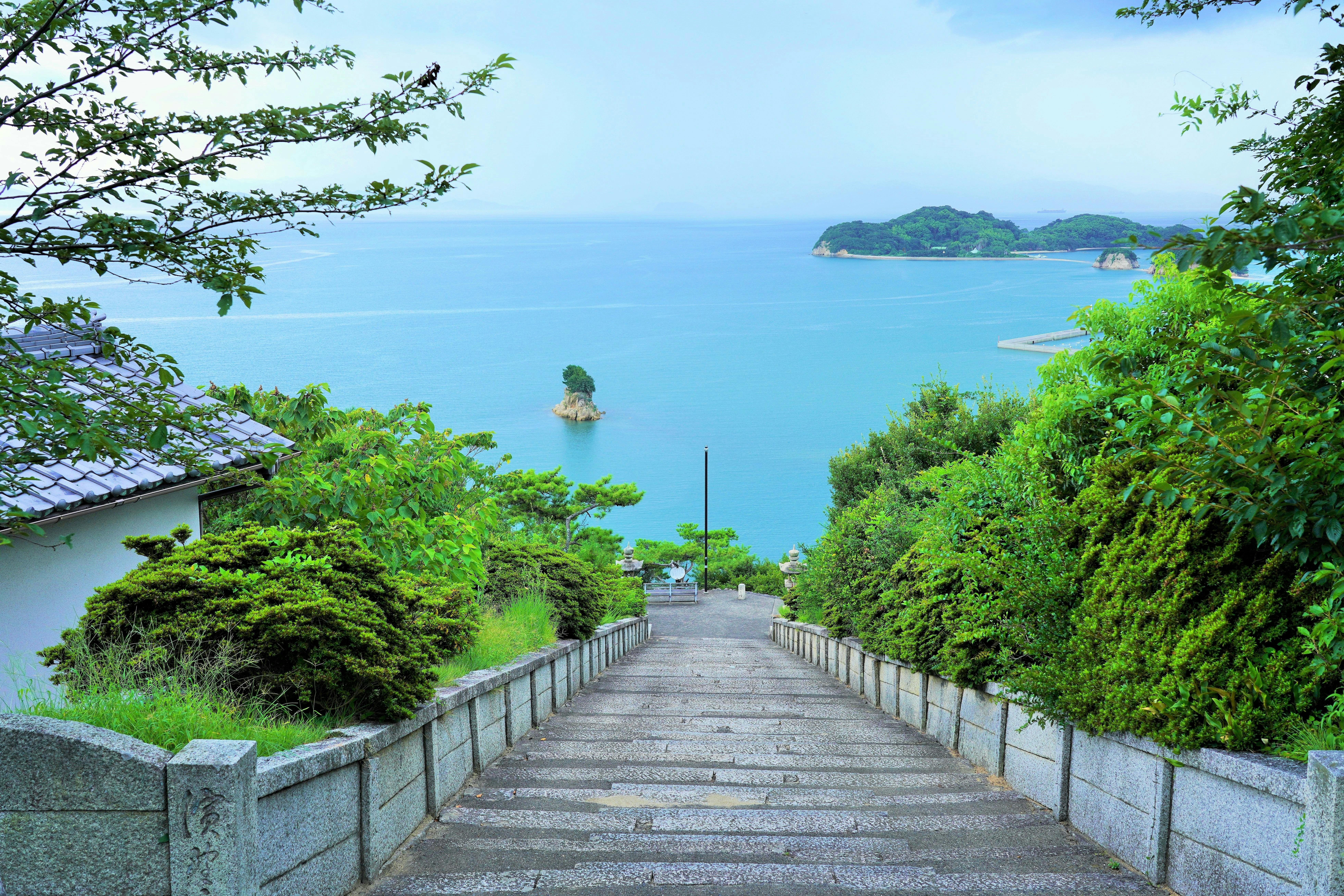 A stone staircase lined with green trees descends toward a calm blue sea, with a small island visible near the shore and larger islands in the distance under a partly cloudy sky.