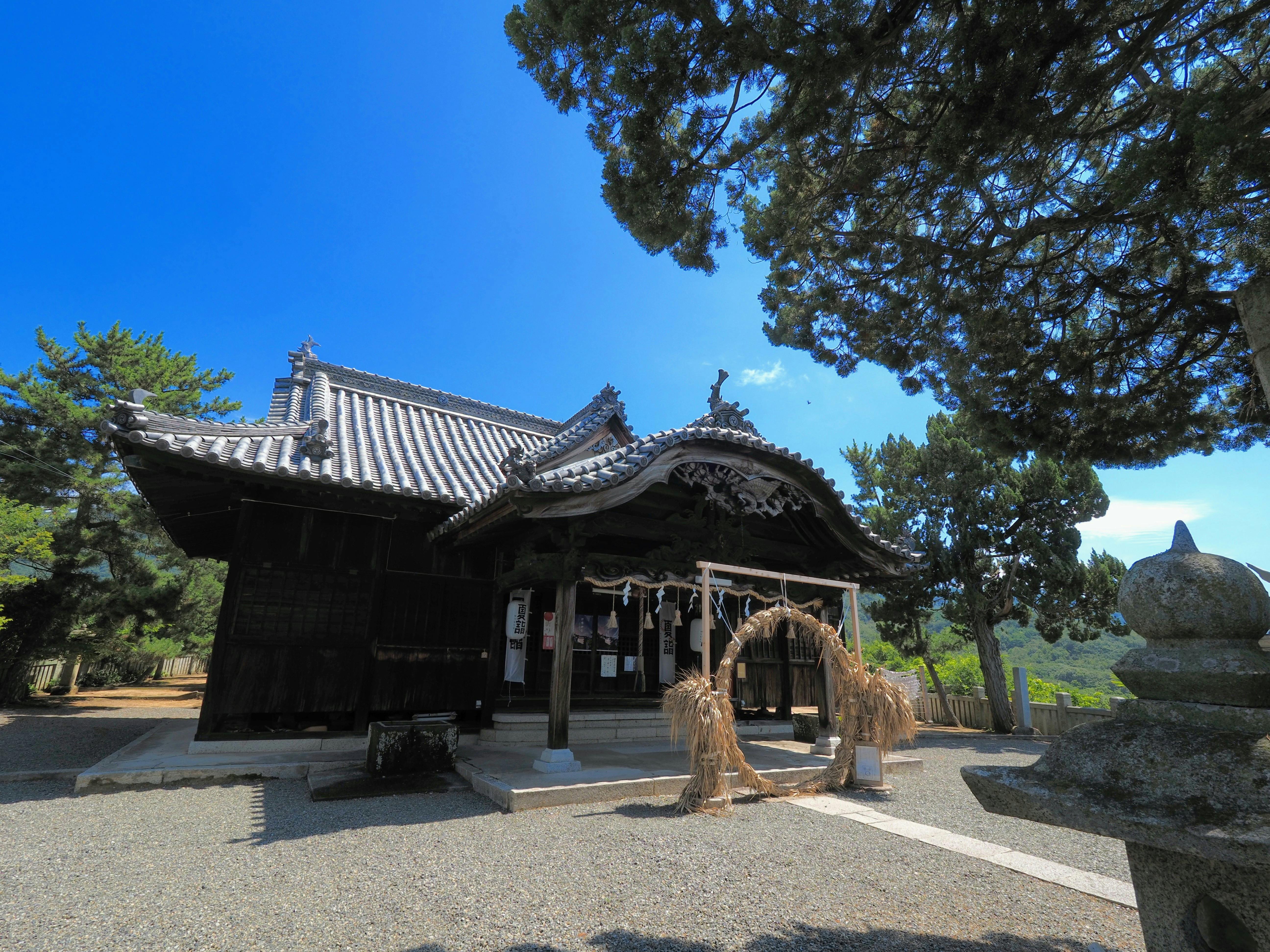 Traditional Japanese shrine with a curved tiled roof, wooden structure, and decorative archway made of straw. Surrounded by trees and bathed in bright sunlight under a clear blue sky.