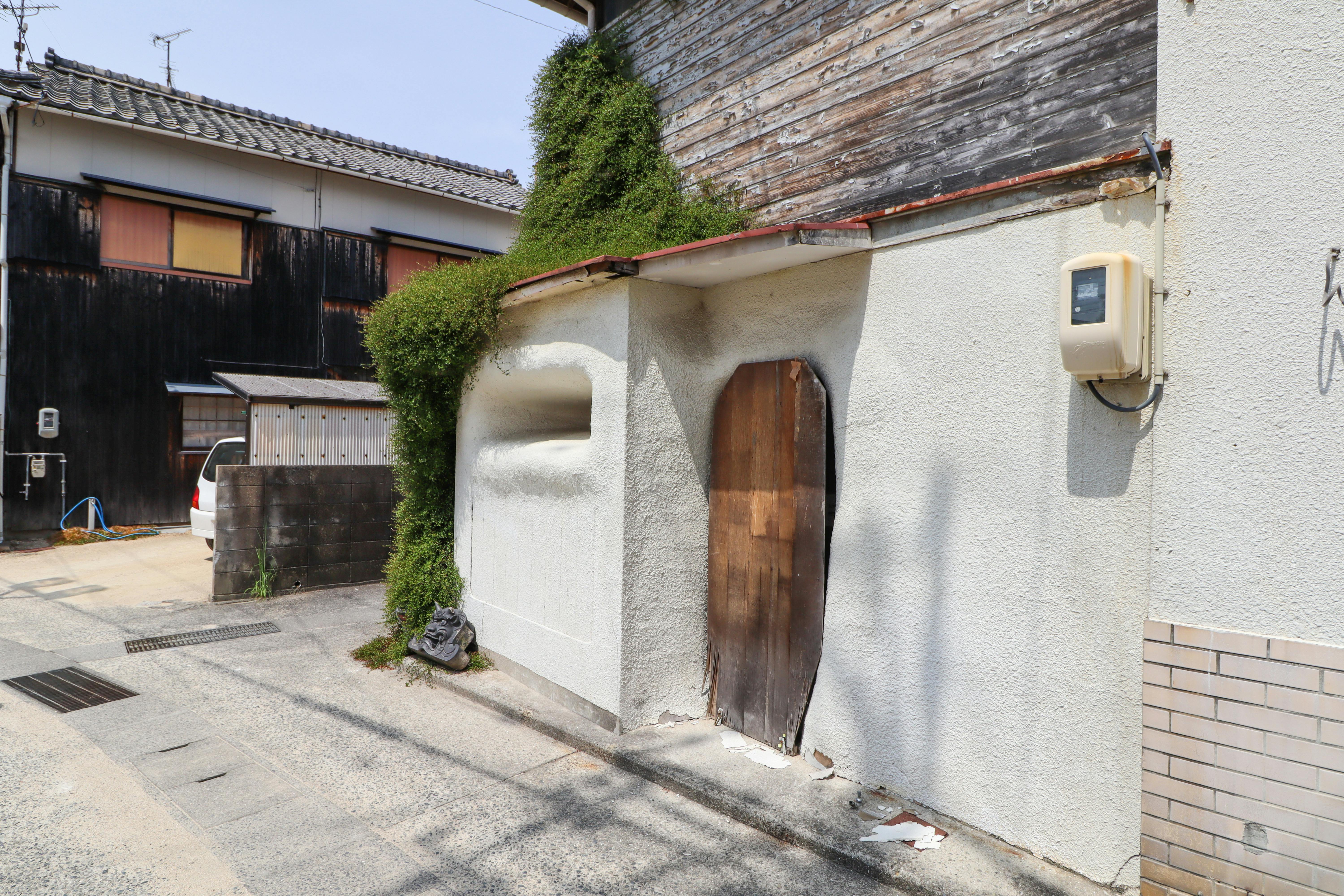 A weathered wooden door set in a white stucco wall with overgrown green vines, next to a utility box, along a quiet residential street with traditional Japanese buildings.