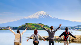 Four people stand with arms raised, facing a serene lake and a snow-capped Mount Fuji in the distance under a clear blue sky. Green trees and a small island are visible in front of the mountain.