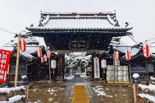 A traditional Japanese temple gate decorated with red lanterns, surrounded by snow-covered rooftops and trees. Barrels are stacked on one side, and snowflakes are gently falling.