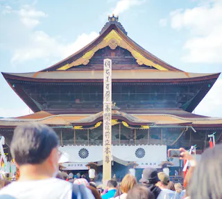 A crowd gathers in front of a large traditional Japanese temple with a wooden facade, gold accents, and white banners. A tall wooden pillar with vertical Japanese writing stands in the center.