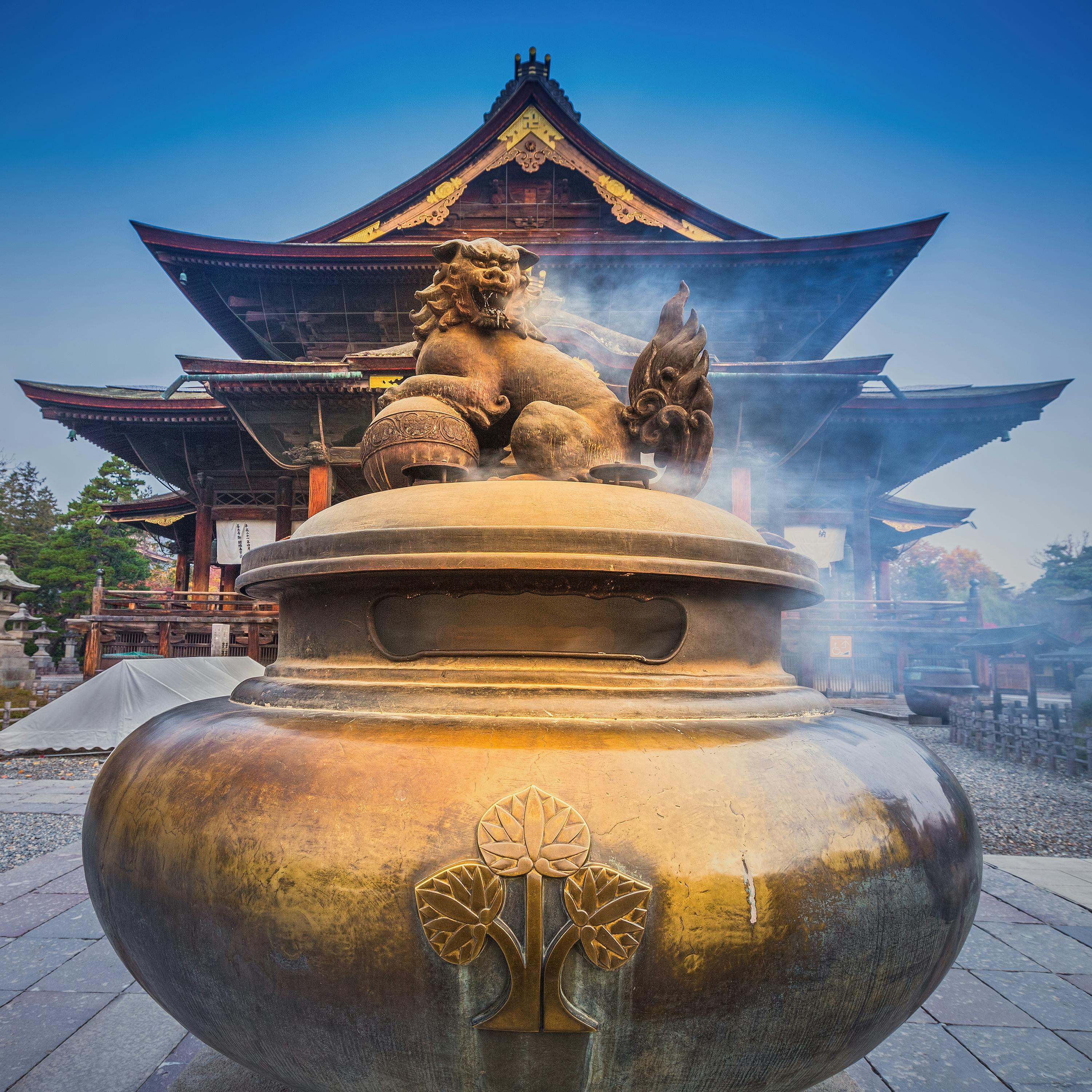 Large bronze incense burner with a lion statue on top emits smoke in front of a traditional Japanese temple with ornate wooden architecture and a sloped roof, under a clear blue sky.