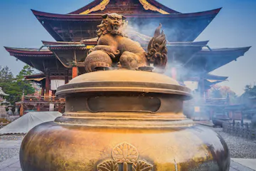 Zenkoji Temple Large bronze incense burner with a lion statue on top emits smoke in front of a traditional Japanese temple with ornate wooden architecture and a sloped roof, under a clear blue sky.