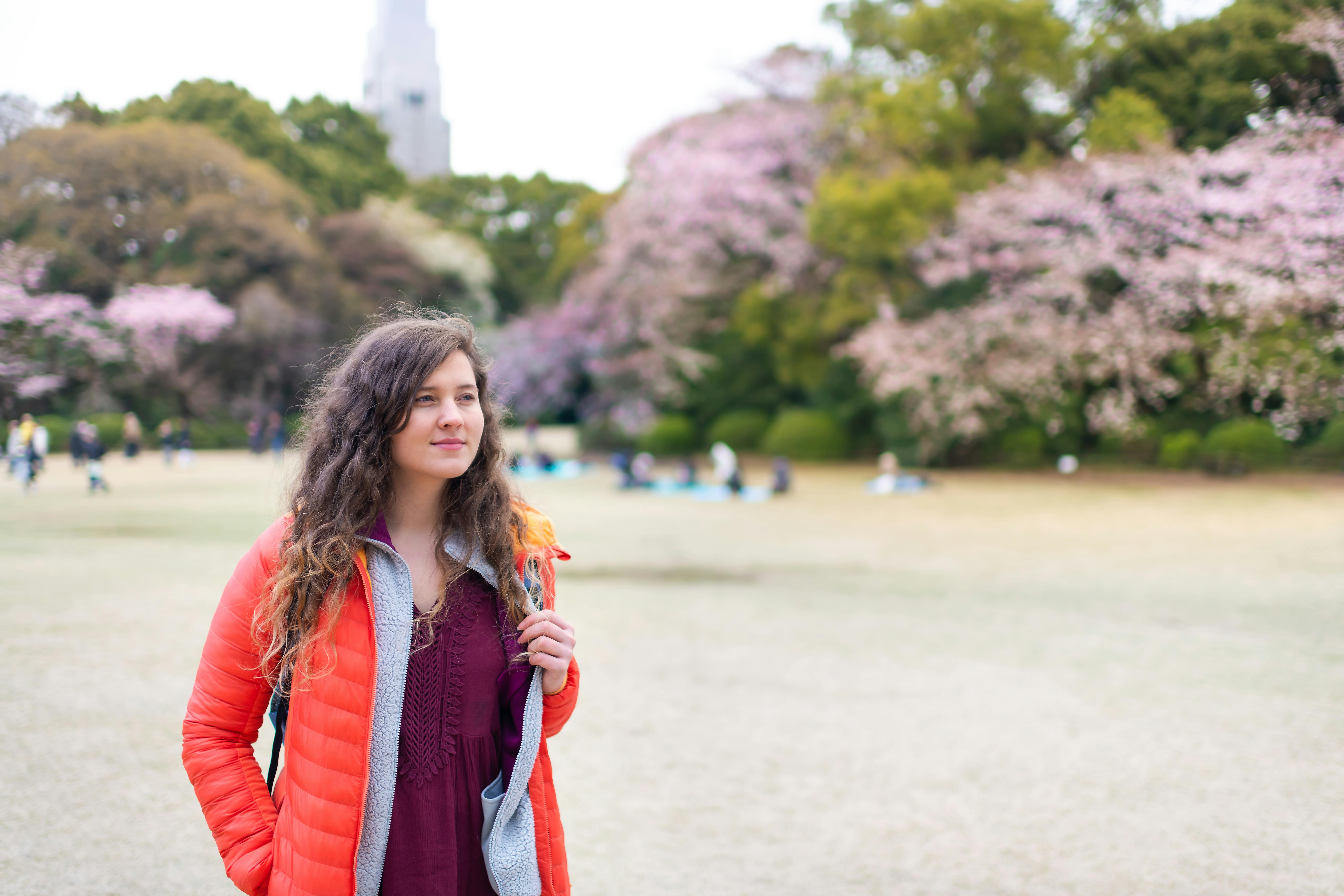 A woman with wavy brown hair wearing an orange jacket stands in a park with cherry blossom trees in bloom. People are sitting on the grass in the background under a cloudy sky.
