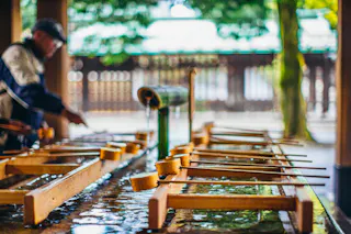 A row of wooden ladles rests on a traditional purification fountain at a Japanese shrine, with a person in the background performing a ritual cleansing. The scene is set outdoors with greenery visible.