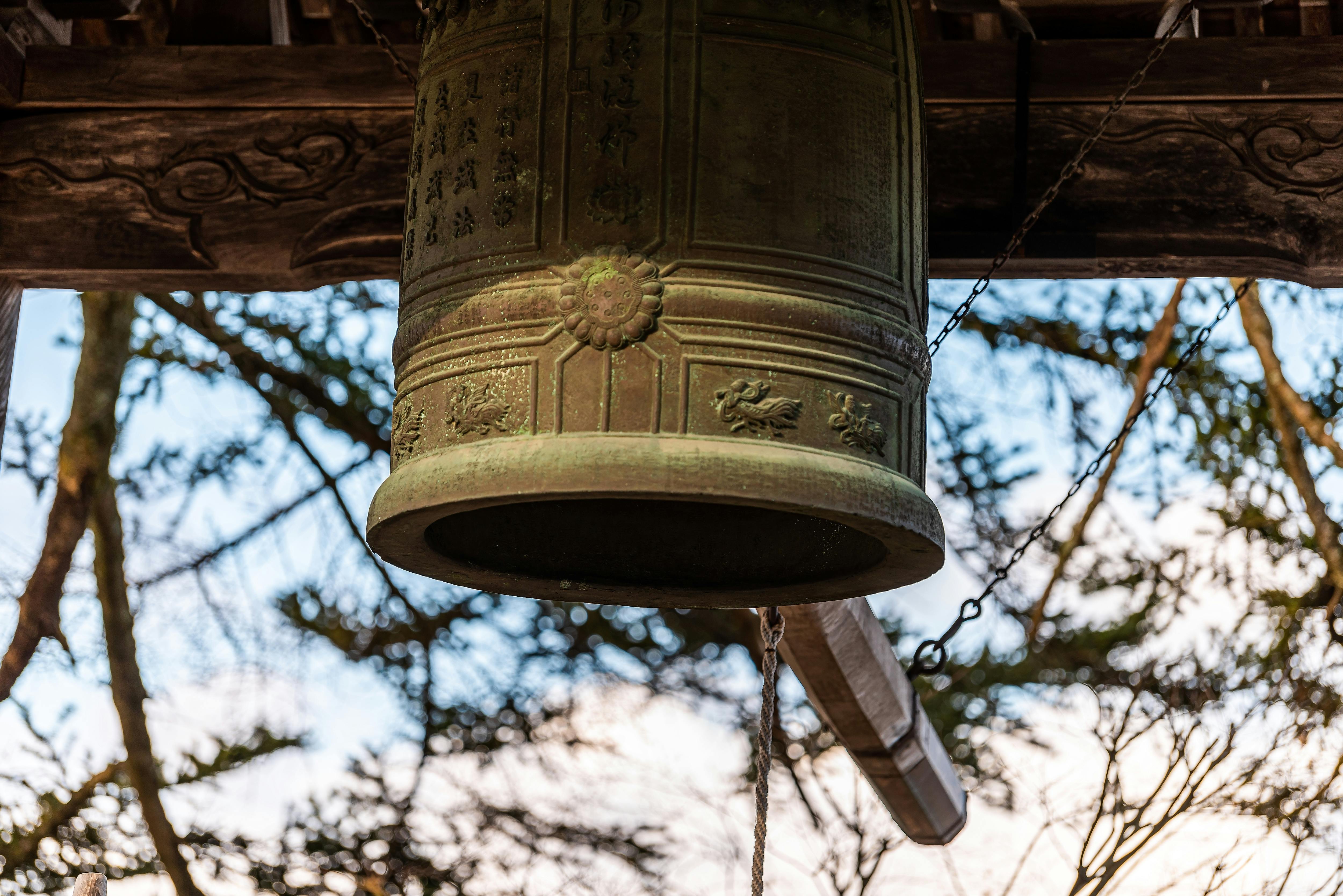 A large, ornate bronze bell with engraved patterns and writing hangs outdoors, with a wooden log suspended beside it for striking. Tree branches and a blue sky are visible in the background.