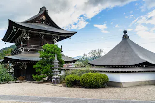A traditional Japanese temple with ornate wooden rooftops, a small pagoda sculpture, green shrubs, and a gravel courtyard under a partly cloudy sky.